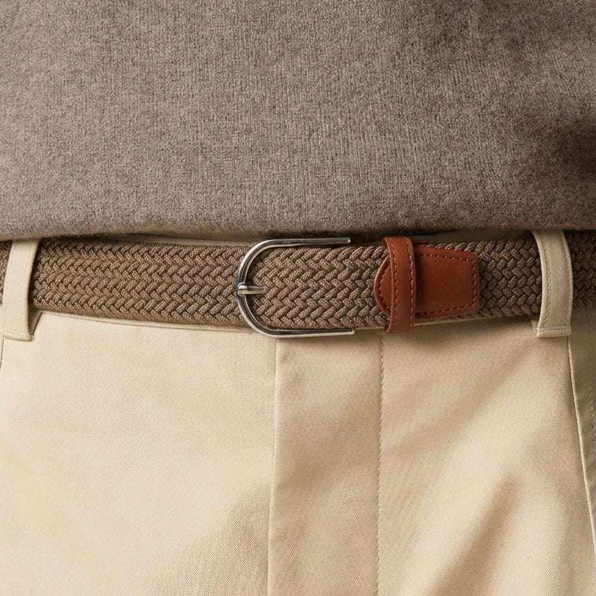 A close-up of a woven mens belt with a silver-toned metal buckle and brown leather tab, worn under a taupe sweater and paired with light beige trousers
