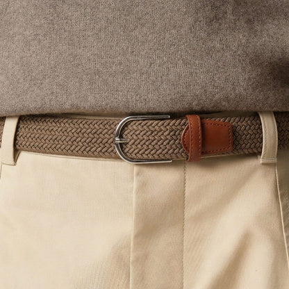 A close-up of a woven mens belt with a silver-toned metal buckle and brown leather tab, worn under a taupe sweater and paired with light beige trousers