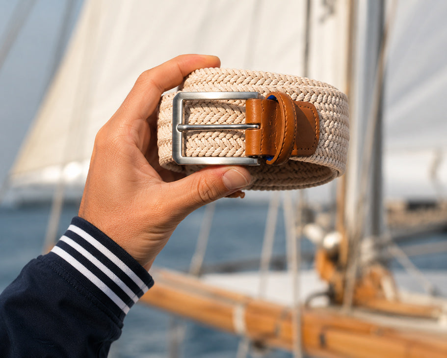 A hand holds a woven beige mens belt with a silver buckle and brown leather accents, with a sailboat in the background