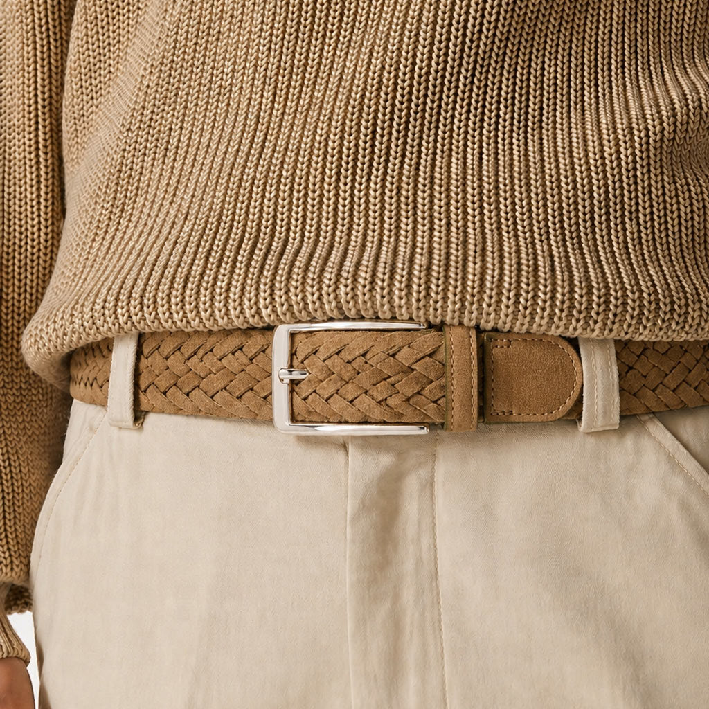 A close-up of a tan woven mens belt with a silver buckle, worn with a beige ribbed sweater and off-white trousers