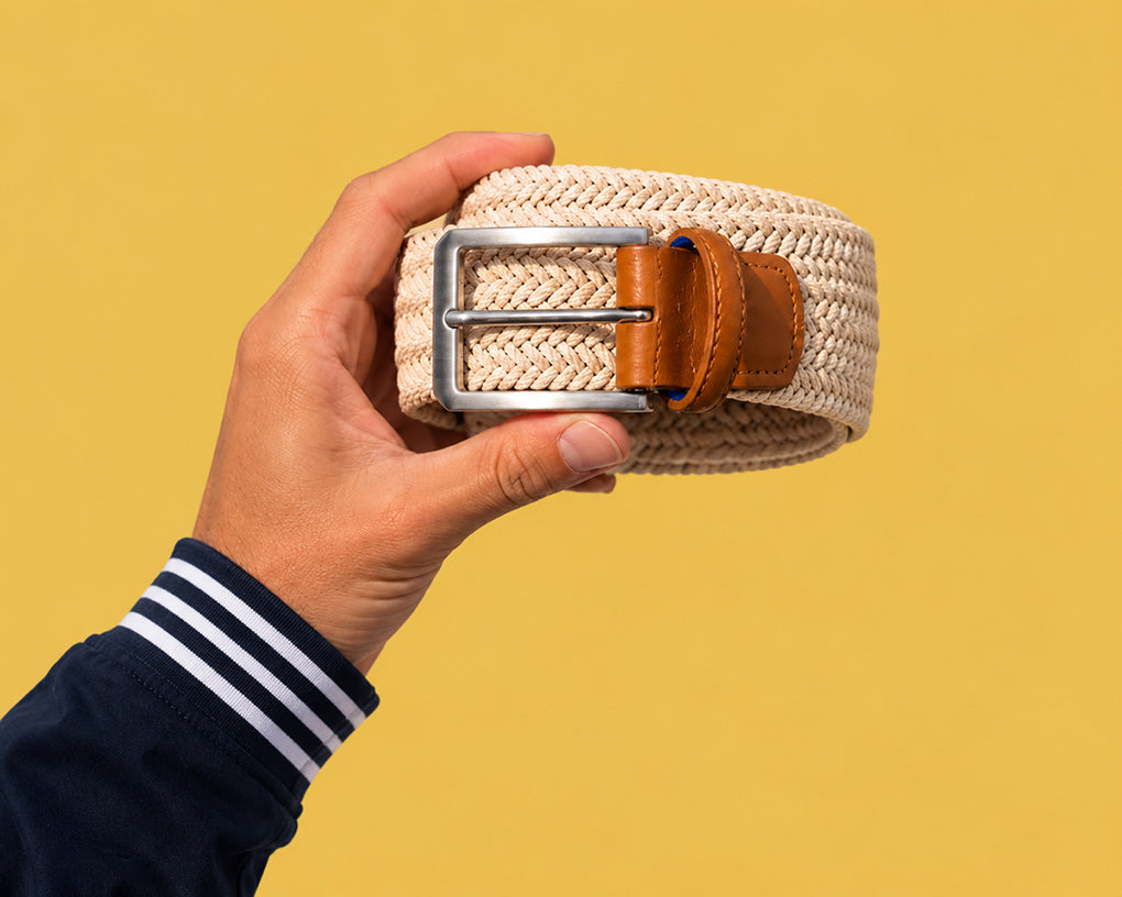 A hand holding a woven beige men's belt with a silver metal buckle and brown leather accents