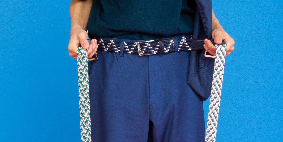 Man wearing a navy woven Beltology belt while holding two additional colorful woven men’s belts against a blue background