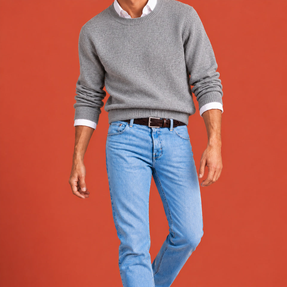 A man wearing a gray crewneck sweater, light blue jeans, a white collared shirt, and a brown men's belt